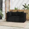 A black poly rattan corner sofa with cushions is placed on a rug on a patio, with plants and wooden fencing in the background. Produktbild-Vorschau