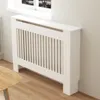 A white radiator cover with vertical slats is shown against a light gray wall, with a window and a wooden table in the foreground. Podgląd zdjęcia produktu
