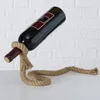 A wine bottle is held up by a rope stand against a white brick wall. Produktbild-Vorschau