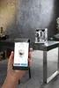 A hand holding a smartphone displaying a coffee machine app, with the actual coffee machine in the background on a counter. Produktbild-Vorschau