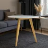 A round white coffee table with wooden legs is placed on a dark gray rug in a living room setting. A gray sofa with a brown cushion is visible to the left, and a floor lamp and curtains are in the background. Podgląd zdjęcia produktu