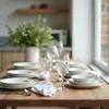 A dining table is set for a meal with plates, glasses, and cutlery, with a vase of greenery in the background. Produktbild-Vorschau