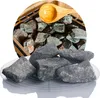 A pile of dark gray sauna stones on a clear plate, with more stones and a wooden scoop visible in the background within a circular frame. Produktbild-Vorschau