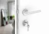 A close-up view of a silver door handle and lock set on a white door, with a blurred interior visible through the open doorway. Produktbild-Vorschau