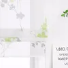 A close-up view of a white roman blind with a green and gray floral pattern, hanging in front of a window. A small green plant is visible on the right side of the image, and a white sign with text is also present. Produktbild-Vorschau