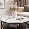 A round coffee table with a white marble top and black base, decorated with a vase of dried flowers, two white taper candles in black holders, a glass carafe, and two glasses on a round tray. A newspaper is visible on the lower shelf. Produktbild-Vorschau