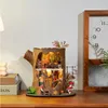 A miniature fairytale tree house model is displayed on a wooden shelf next to a clock and some books. Podgląd zdjęcia produktu