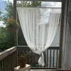 A white sheer curtain is hanging on a wooden balcony, with plants in the background. Produktbild-Vorschau