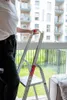 A person is standing next to a ladder in front of a window with a view of a building and greenery. Anteprima dell’immagine prodotto