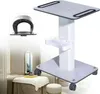 A white rolling trolley with a tray and a top shelf, shown in a salon-like setting with a massage table and a mirror in the background. A smaller inset image shows a close-up of a beauty device. Náhled obrázku produktu