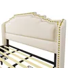 A close-up view of a beige upholstered bed frame with gold nailhead trim. Náhled obrázku produktu