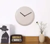 A minimalist wall clock with black hands is mounted on a white wall above a wooden shelf. On the shelf, there is a vintage-style radio and a vase with dried flowers. To the left of the clock, a book with a black and white image of a person wearing a hat is Voir les images du produit