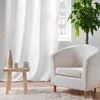 A minimalist living room scene with a white armchair, a small wooden side table with decorative items, and a large white curtain. Produktbild-Vorschau