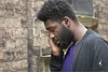 A man wearing earbuds leans against a brick wall. Anteprima dell’immagine prodotto