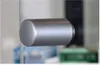 A close-up shot of a silver cylindrical handle. The background is blurred and shows elements of what appears to be a glass door or window frame with some electronic components visible in the background. Anteprima dell’immagine prodotto