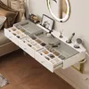 A white vanity table with glass top and drawers filled with makeup, placed next to a bed and a wall with a round mirror. Voir les images du produit