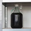 A glass carboy filled with dark liquid sits on a shelf, with a fermentation airlock attached to the top. Produktbild-Vorschau