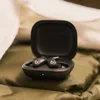 Black wireless earbuds in an open charging case, placed on a green textured surface with a white cloth in the background. Produktbild-Vorschau