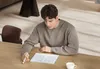 A man is sitting at a wooden table, using a stylus to write on a tablet. Produktbild-Vorschau