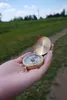 A hand holding a compass in a grassy outdoor setting. Produktbild-Vorschau