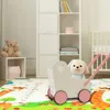 A nursery room scene with a play mat, a teddy bear, a toy stroller with a plush dog, and a crib in the background. Produktbild-Vorschau