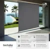 A grey vertical awning is shown deployed in front of a large glass sliding door, with a scenic view of a beach and ocean visible through the door. Text and icons describing the product's features are displayed in the lower right corner. Náhled obrázku produktu