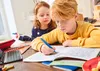 Two children are doing homework at a table. The boy is writing in a notebook with a blue pen, and the girl is looking at his work. Náhľad obrázku produktu