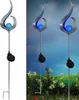 A solar-powered garden stake with a blue orb and a flame-like metal design, shown both on a white background and in a garden setting. Produktbild-Vorschau