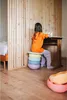 A young girl sits at a desk, using a stack of colorful cushions as a stool. The scene is set in a room with wooden paneling and a bed visible in the background. Anteprima dell’immagine prodotto
