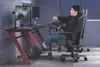 A man playing a racing simulator game on a gaming desk with multiple monitors, a racing wheel, pedals, and a gaming chair. Produktbild-Vorschau
