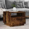 A wooden side table with a drawer and an open shelf, placed in a living room setting next to a grey sofa with cushions and a throw blanket. Produktbild-Vorschau