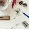 A flat lay of art supplies including a partially drawn flower, pencils, erasers, and pencil shavings. Produktbild-Vorschau
