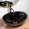 A black and gold marble-patterned oval sink with a gold faucet, placed on a wooden surface, with a gold-rimmed mirror in the background. Náhľad obrázku produktu