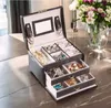 A black jewelry box with multiple compartments and drawers, filled with various jewelry items, sits on a marble countertop next to a vase of flowers. Náhľad obrázku produktu
