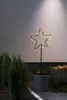 A lighted star decoration in a pot with evergreen branches and red ornaments, placed outdoors next to evergreen bushes and a basket of firewood. Podgląd zdjęcia produktu
