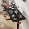 A black standing desk with laptops, a keyboard, and a mouse on it, set up in a home office environment with a plant and a window in the background. Voir les images du produit