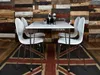 A white dining table with four white chairs is set up on a Union Jack rug in front of a rustic wooden wall. Podgląd zdjęcia produktu