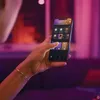 A person is holding a smartphone, interacting with a smart lighting app. The background is illuminated with purple and pink ambient light. Produktbild-Vorschau