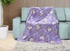 A purple unicorn-themed blanket is draped over a light blue couch in a living room setting. Náhled obrázku produktu