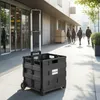 A black folding shopping trolley with a telescopic handle stands on a paved sidewalk in front of a modern building with large glass windows. Several people are walking in the background. Náhled obrázku produktu