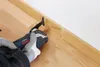 A person wearing gloves is using an oscillating multi-tool to cut a wooden baseboard near a wall. Produktbild-Vorschau