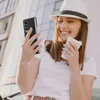 A woman in a white hat and t-shirt is holding a phone and a coffee cup, smiling. Náhled obrázku produktu