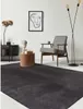 A dark gray rug is placed in a modern living room setting, in front of a wooden side table with a bust and books, and a patterned armchair. Produktbild-Vorschau