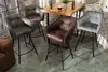 Four bar stools are arranged around a wooden table in a room with a brick wall. Produktbild-Vorschau