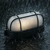 A black outdoor light fixture with a ribbed white diffuser is shown in heavy rain, with water splashing around it and pooling on the surface it sits on. Produktbild-Vorschau