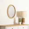 An oval mirror with a gold frame hangs on a white wall above a wooden dresser. A table lamp, a potted plant, and a candle are on the dresser. Náhľad obrázku produktu