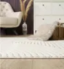 A close-up view of a cream-colored rug with a geometric pattern, placed on a wooden floor in a room with a chair, a vase with dried grass, and a storage unit in the background. Produktbild-Vorschau