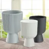 Three modern planters of different sizes and colors (gray, white, and black) are displayed on a wooden surface in front of a blurred window with greenery outside. Podgląd zdjęcia produktu