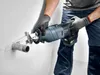 A person wearing gloves and work clothes is using a cordless reciprocating saw to cut a pipe protruding from a wall. Voir les images du produit