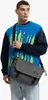 A man wearing a blue and navy sweatshirt with a graphic print, carrying a gray messenger bag. Anteprima dell’immagine prodotto